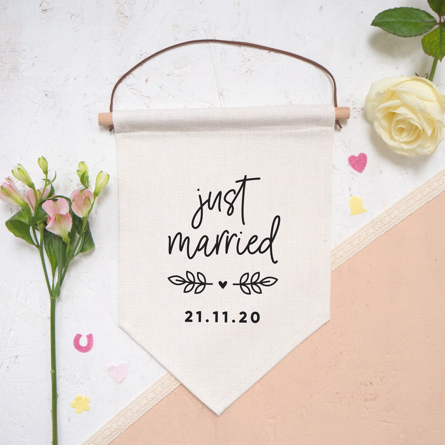 Just married personalised pennant flag with script 'just married', vine and a heart detail and a personalised wedding date. The flag is photographed on a white and peach background with confetti and flowers.