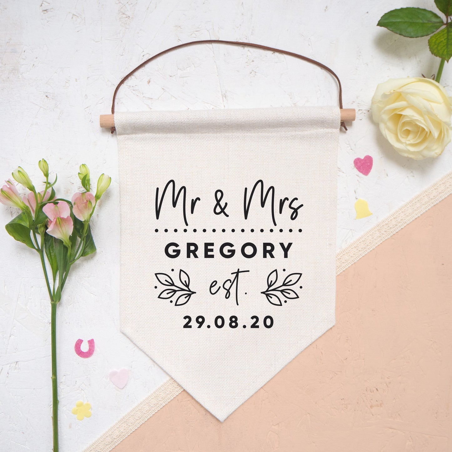 A personalised wedding pennant flag featuring your new title e.g. Mr & Mrs, your new family name and the date that you tied the knot. Photographed on a white and peach background with confetti and flowers either side of the flag.