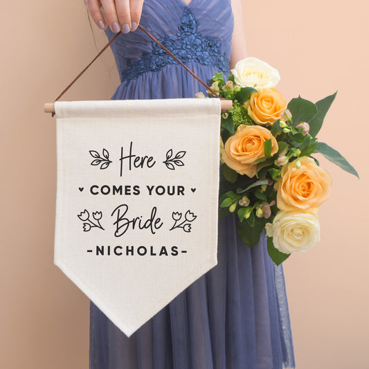 A linen pennant flag with the words 'here comes your bride [insert name of groom]. Each flag features leafy vines and little hearts. This flag has been photographed with a bridesmaid in the background wearing a purple dress and holding a peach bouquet of flowers.