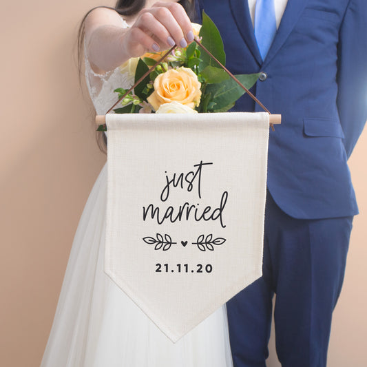 Just married personalised pennant flag with script 'just married', vine and a heart detail and a personalised wedding date. The flag is held by the bride and stood with the groom in a blue suit.