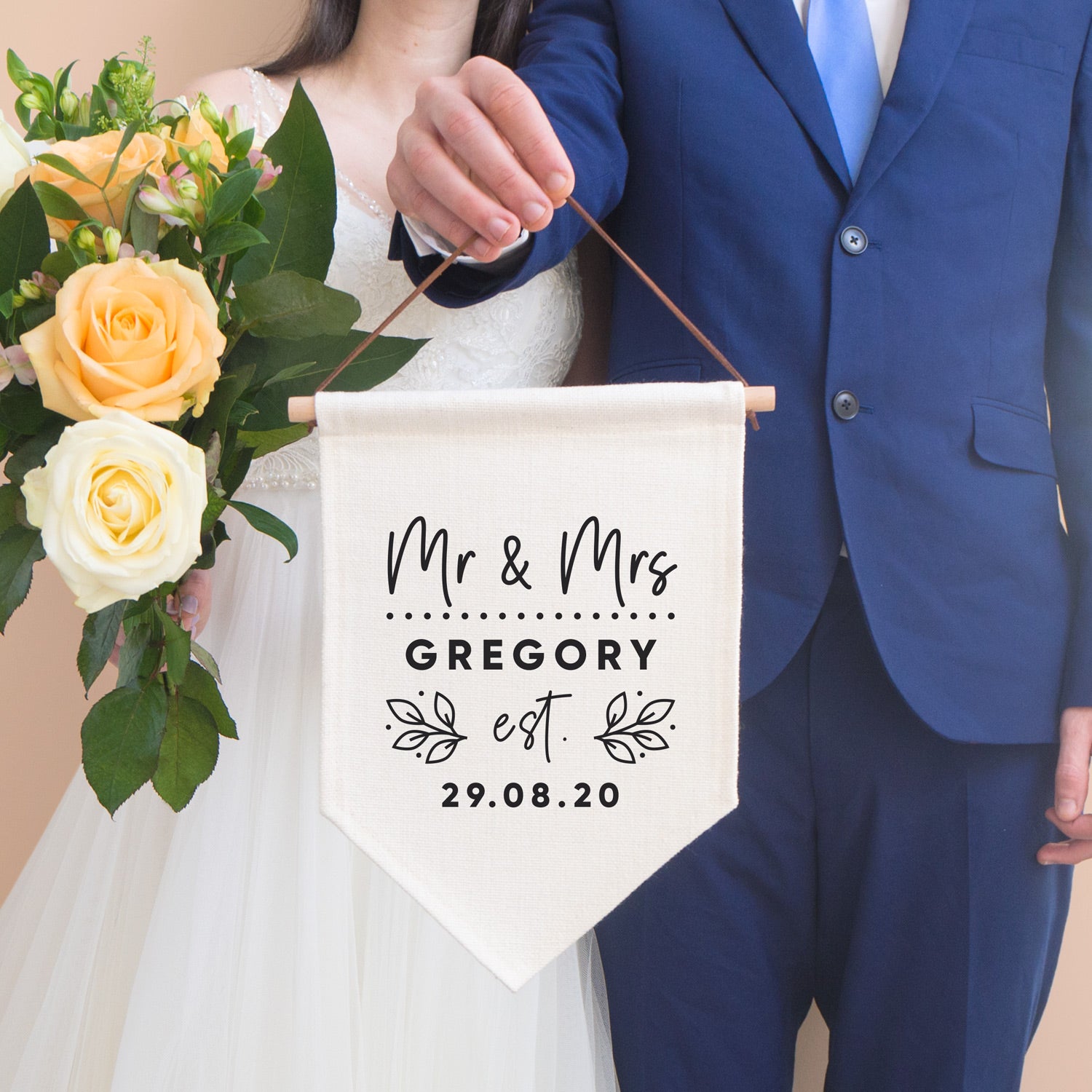 A personalised wedding pennant flag featuring your new title e.g. Mr & Mrs, your new family name and the date that you tied the knot. This linen flag is being held by the groom in a blue suit and the bride is to the left holding a bouquet of peach roses.