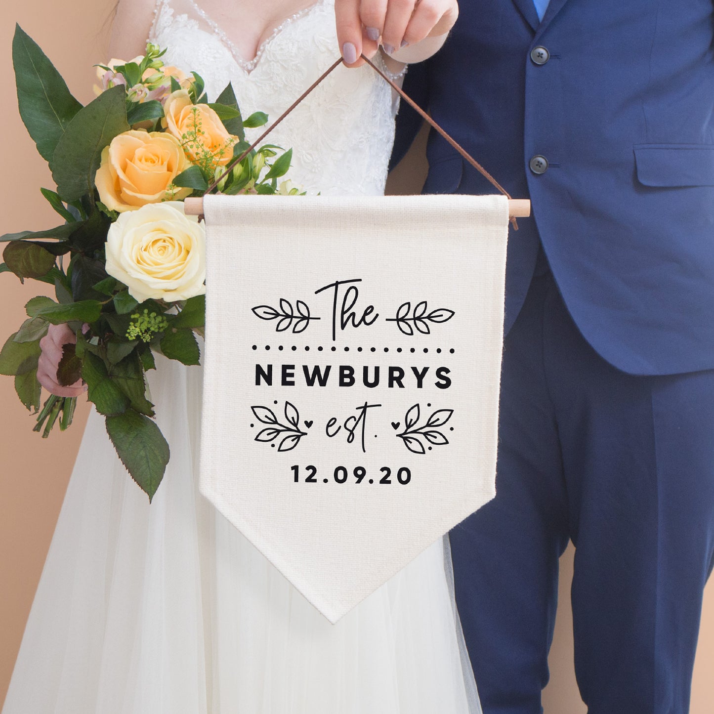 A personalised wedding pennant flag that reads' The [family name] est. [wedding date]. The text is surrounded by leafy vines and love hearts. A bride is holding the flag with a bouquet of peach flowers and is stood next to a groom in a blue suit.