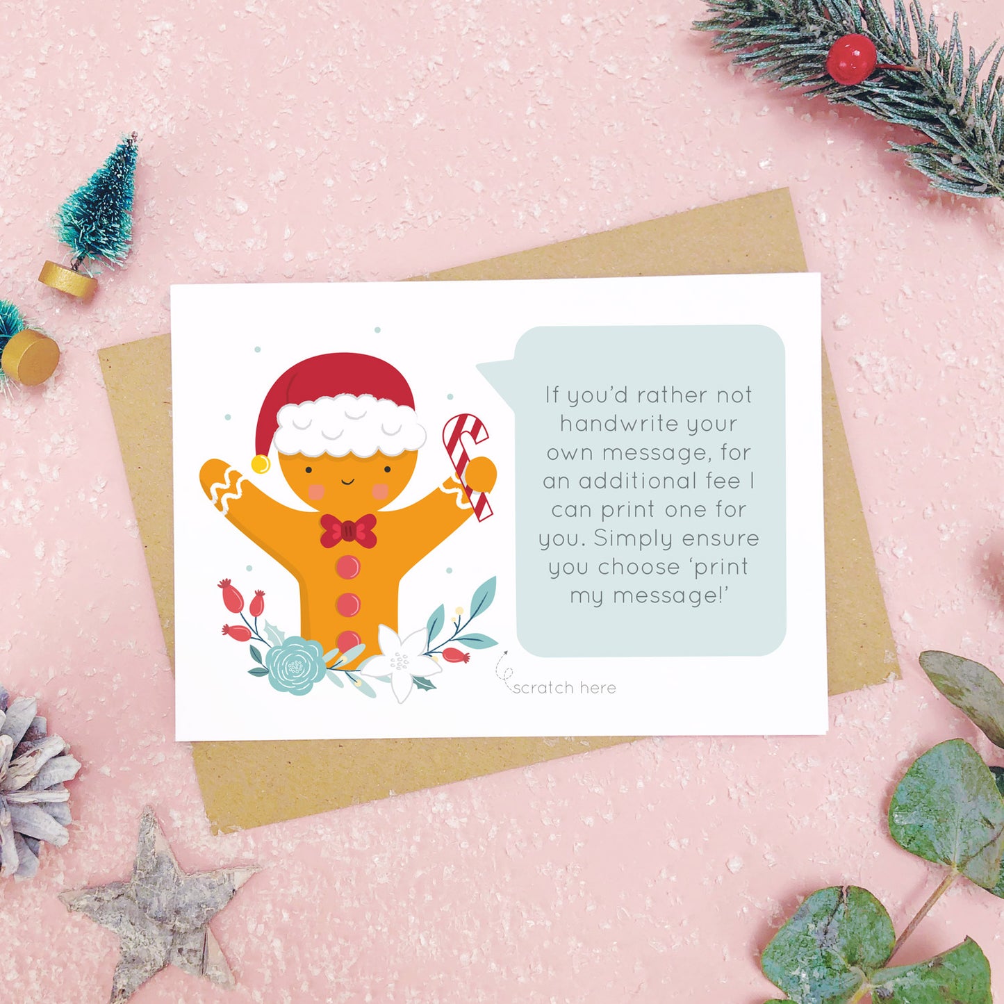 A personalised gingerbread man scratch card an example of the printed message. Shot on a pink background with grey and green festive props.