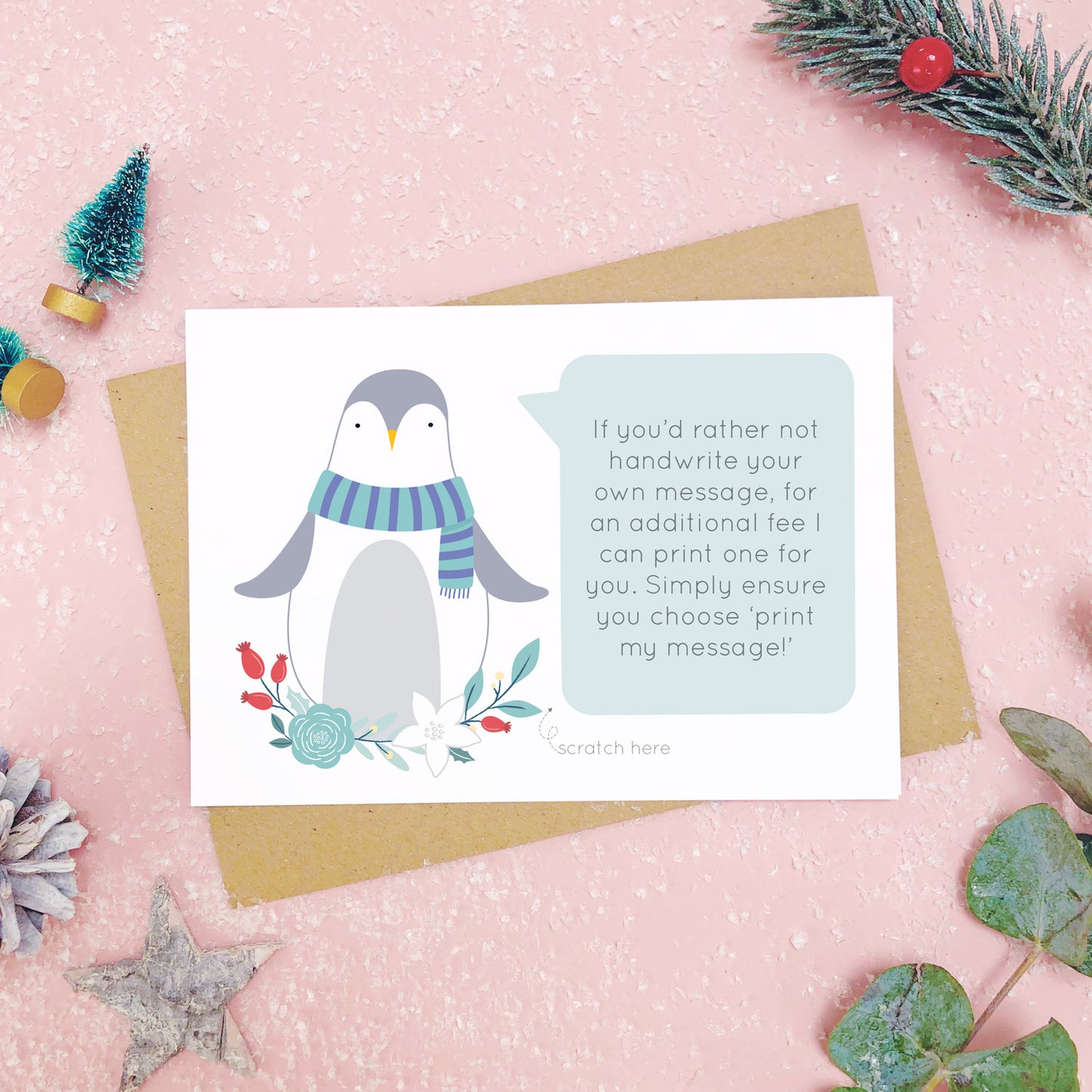 Penguin scratch card showing where you write your hidden message can be printed. Shot on a pink background, surrounded with festive christmas props in tones of green and grey.