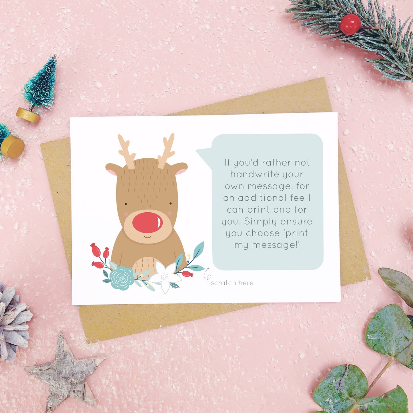 A personalised red nose reindeer scratch card an example of the printed message. Shot on a pink background with grey and green festive props.