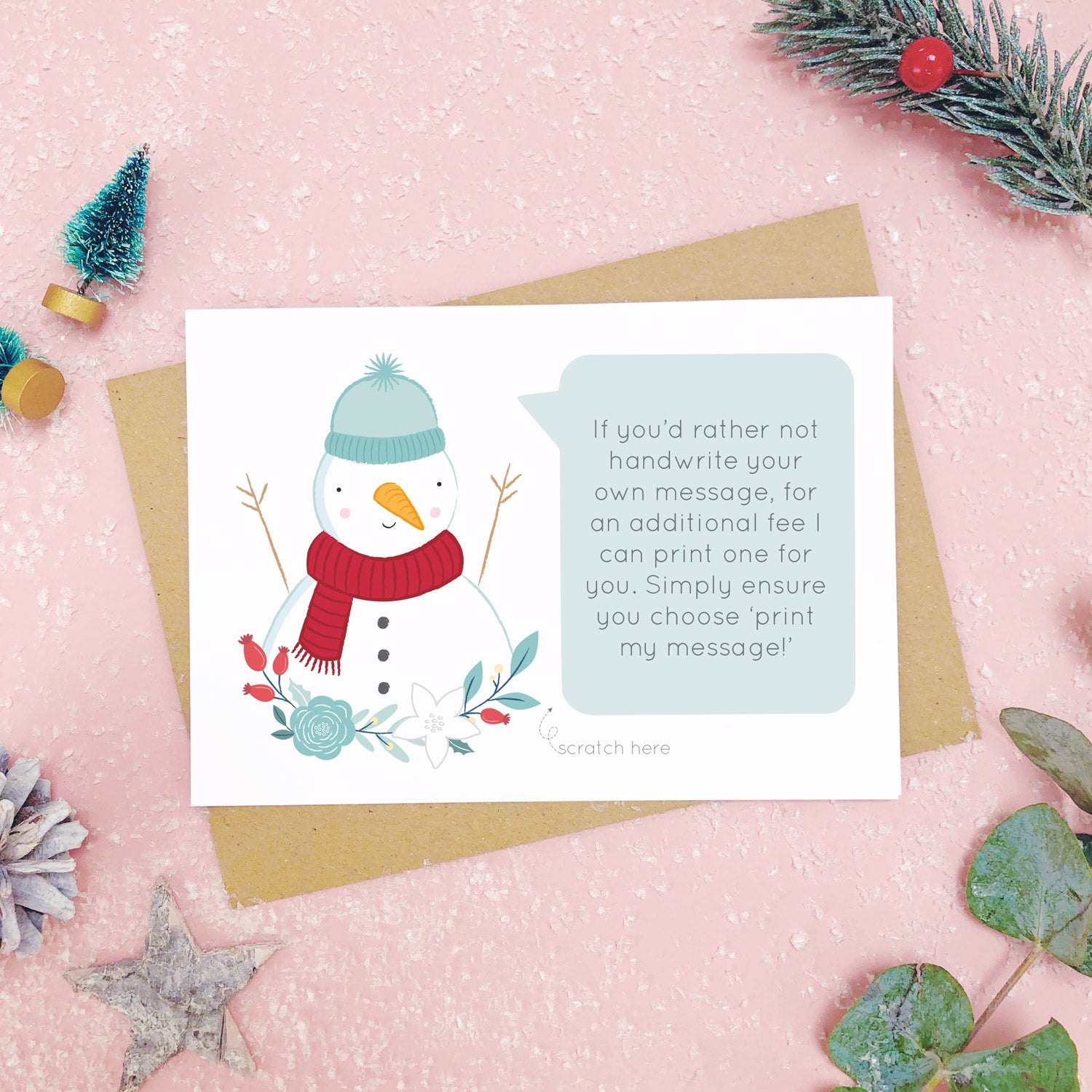 A personalised snowman scratch card an example of the printed message. Shot on a pink background with grey and green festive props.