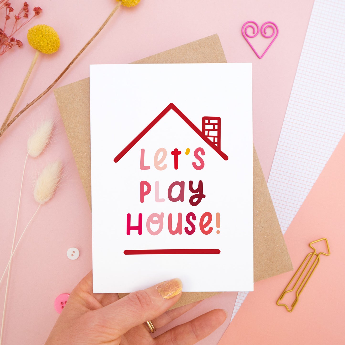 The 'let's play house' card photographed on a pink background with dried flowers, buttons and paper clips as props. The card itself is being held above the scene.