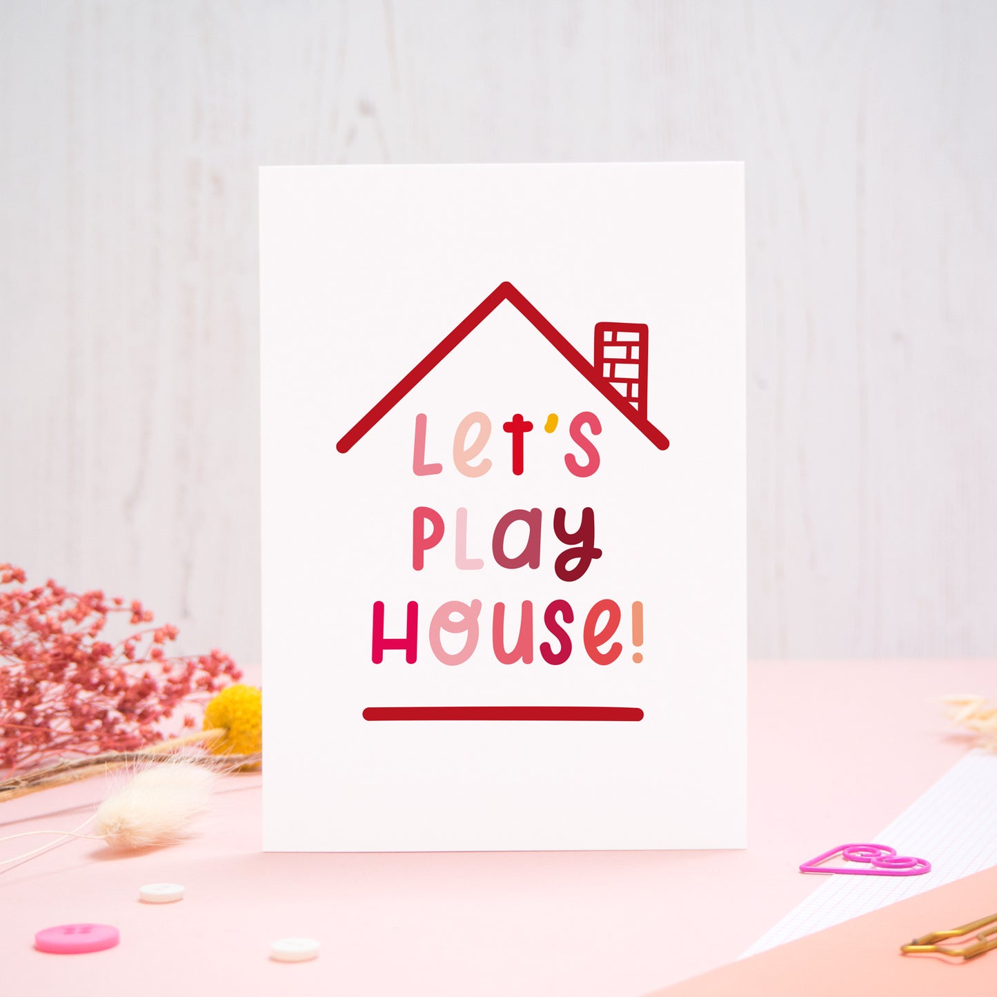 The 'let's play house' card photographed standing up against an off white background with flowers, buttons and paperclips at the base of the card.