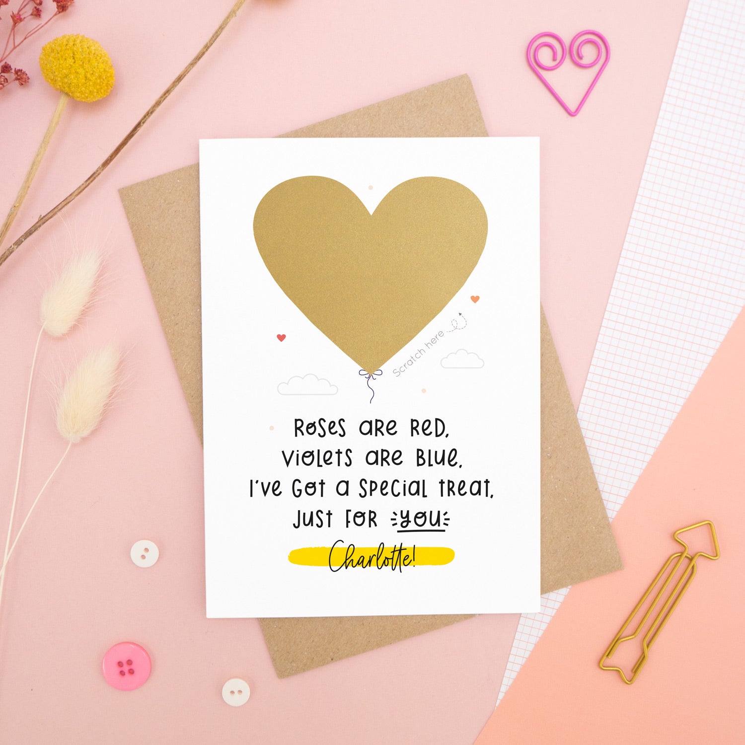 A personalised anniversary scratch card photographed on a pink background with floral props, paper clips, and buttons. This card shows the golden heart before it gets scratched off to reveal the secret message!