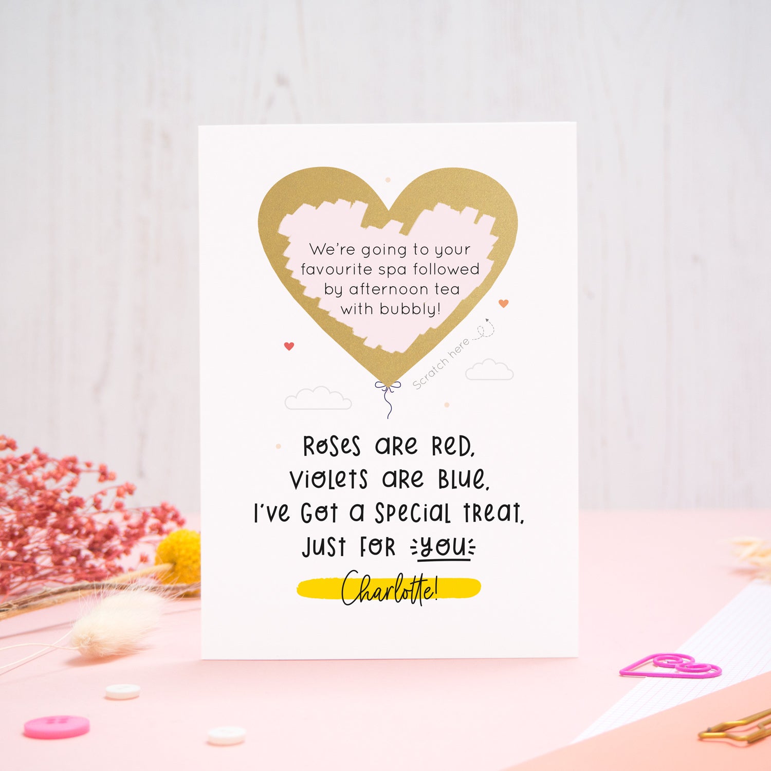 A personalised anniversary scratch card photographed on a pink and white background with floral props, paper clips, and buttons. This card shows the golden heart after it has been scratched off to reveal the secret message!