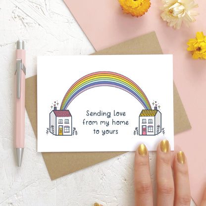 A sending love, rainbow house card photographed on a white and pink background with a pink pen, dried flowers and a hand.. The card features two houses being joined together with a pastel rainbow and text that reads 'sending love from my home to yours'.