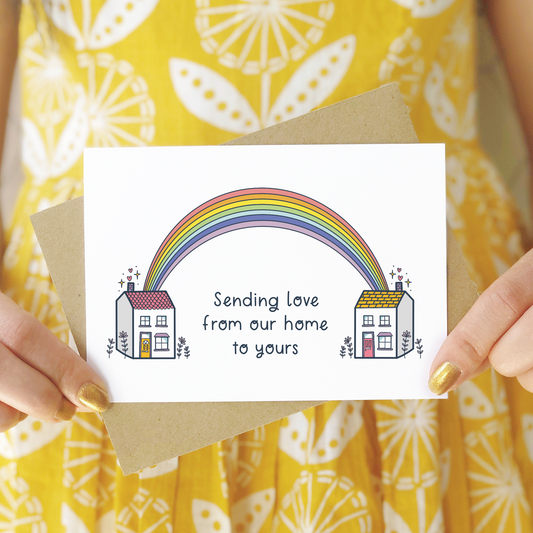 A sending love, rainbow house card photographed being held in front of a person wearing a yellow patterned dress. The card features two houses being joined together with a pastel rainbow and text that reads 'sending love from our home to yours'.