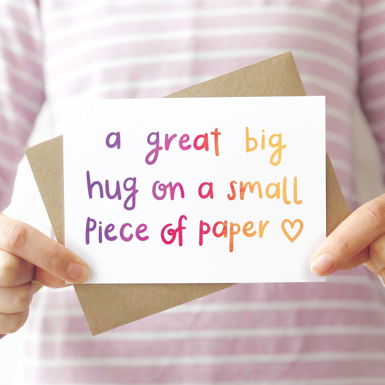 A great big hug on a small piece of paper card photographed being held in front of a pink stripey shirt.