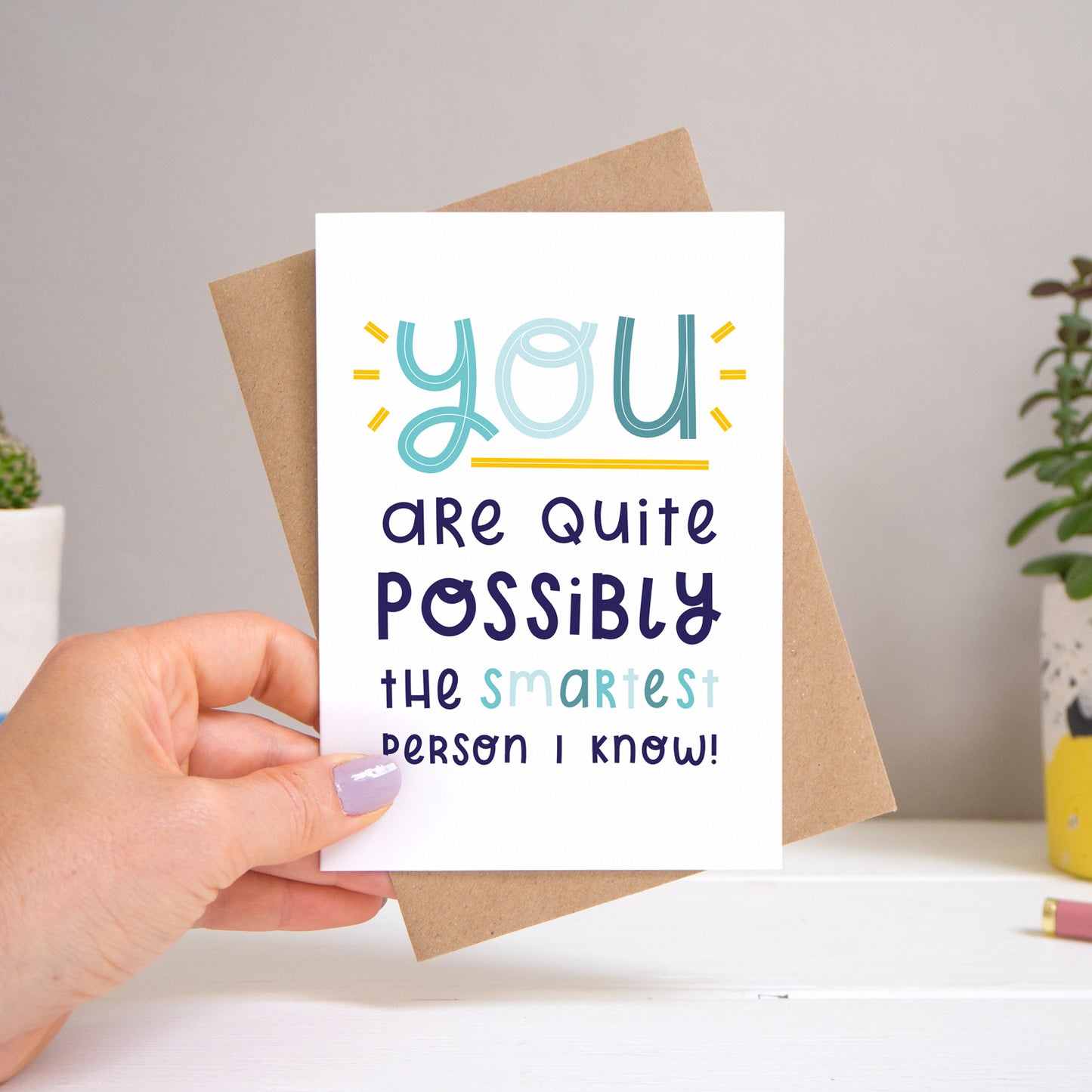 A ‘smartest person I know’ card held over a warm grey and white background with potted plants peeping the sides. Behind the card is a kraft brown envelope that comes with the card. The text on this version of the card is in varying tones of navy and blue.