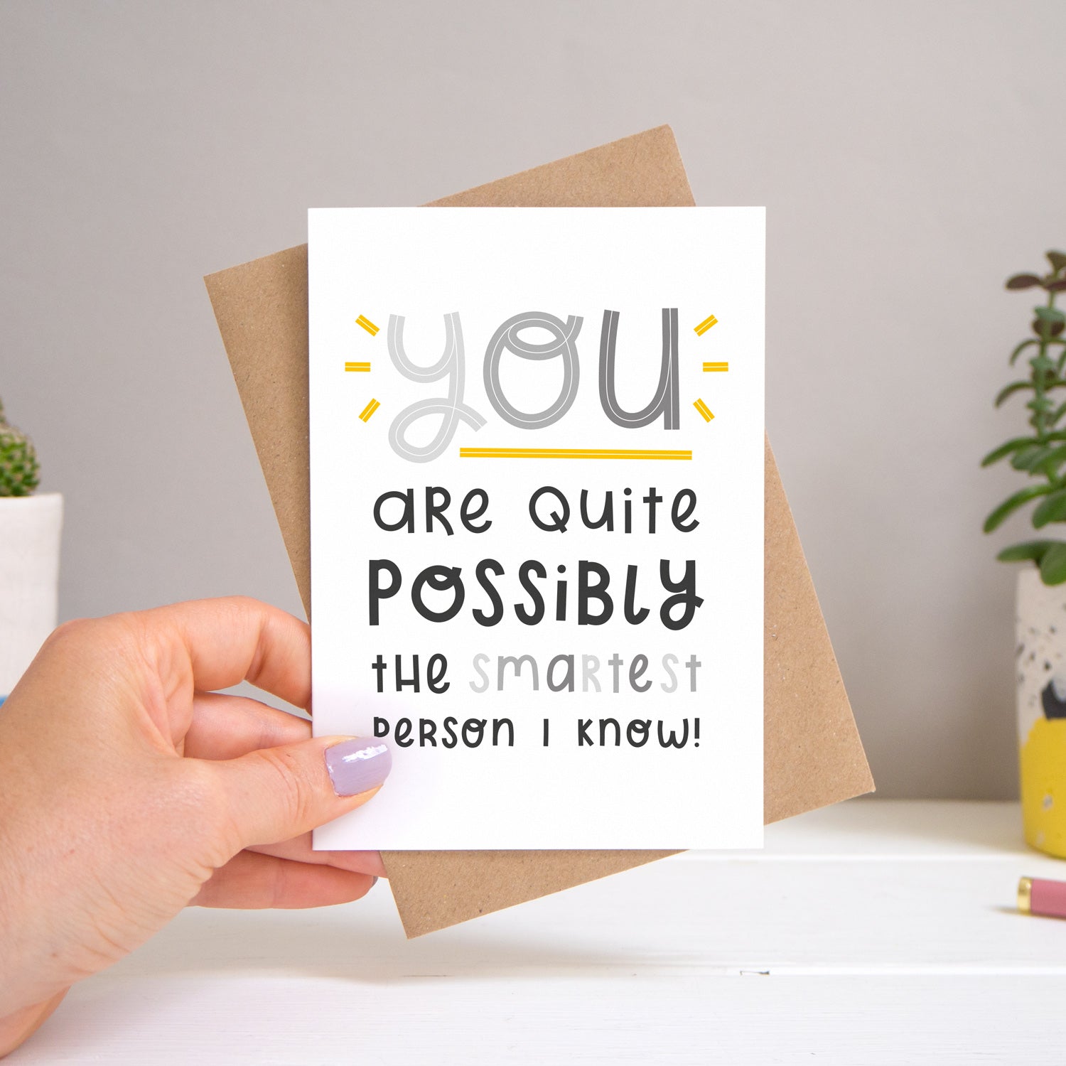 A ‘smartest person I know’ card held over a warm grey and white background with potted plants peeping the sides. Behind the card is a kraft brown envelope that comes with the card. The text on this version of the card is in varying tones of grey.