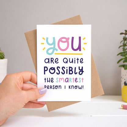 A ‘smartest person I know’ card held over a warm grey and white background with potted plants peeping the sides. Behind the card is a kraft brown envelope that comes with the card. The text on this version of the card is in varying tones of navy and pink, purple and blue.