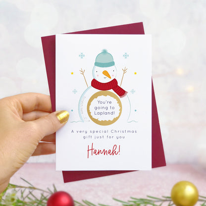 A personalised snowman christmas scratch card photographed being held by a hand to the left and foliage and baubles in the bottom of the frame. The card is against its ‘red wine’ colour envelope and the scratch off panel has been scratched off to reveal a trip to lapland!