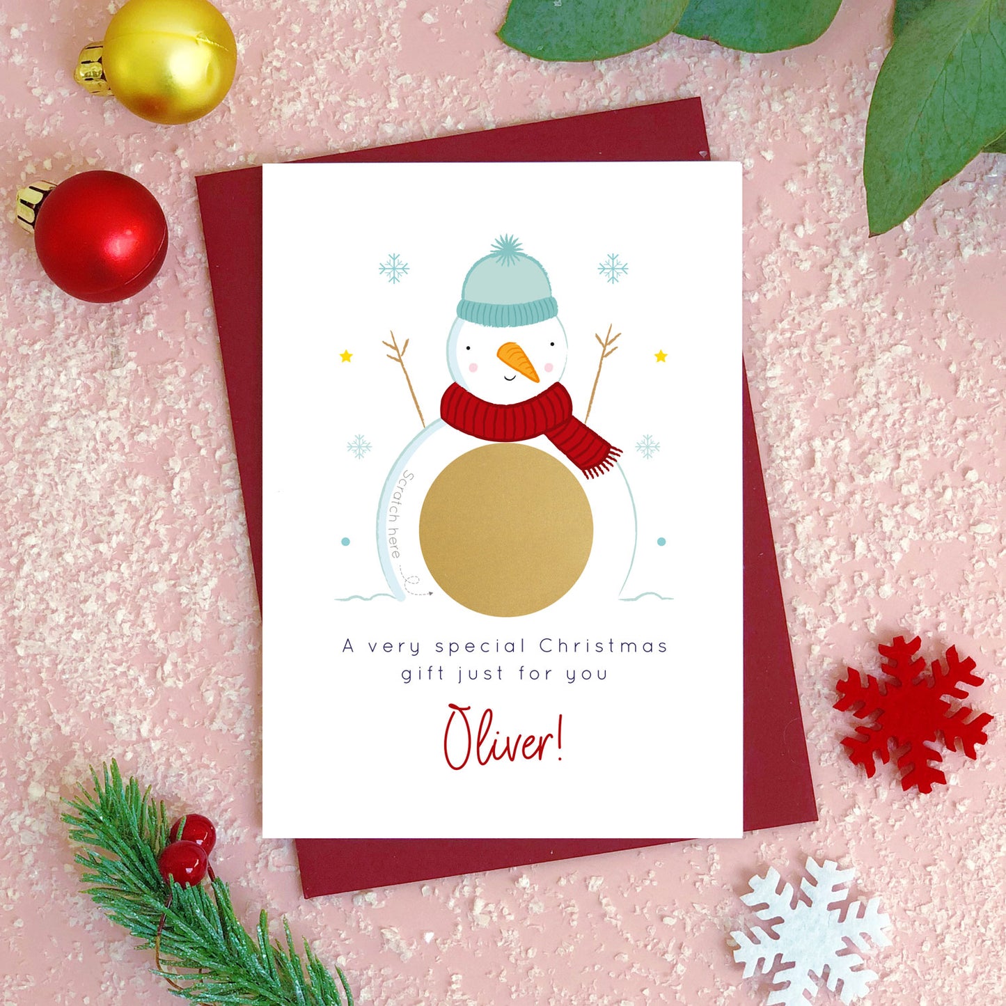 A personalised snowman christmas scratch card photographed flat lying on a red wine coloured envelope on a pink surface surrounded by fake snow, baubles and foliage. The gold panel has not yet been scratched off to reveal the custom message.