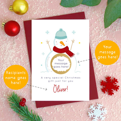A personalised snowman Christmas scratch card photographed flat lying on a red wine coloured envelope. The orange circles demonstrate which areas of the card can be personalised. Here they point to the message and name for the front of the card.