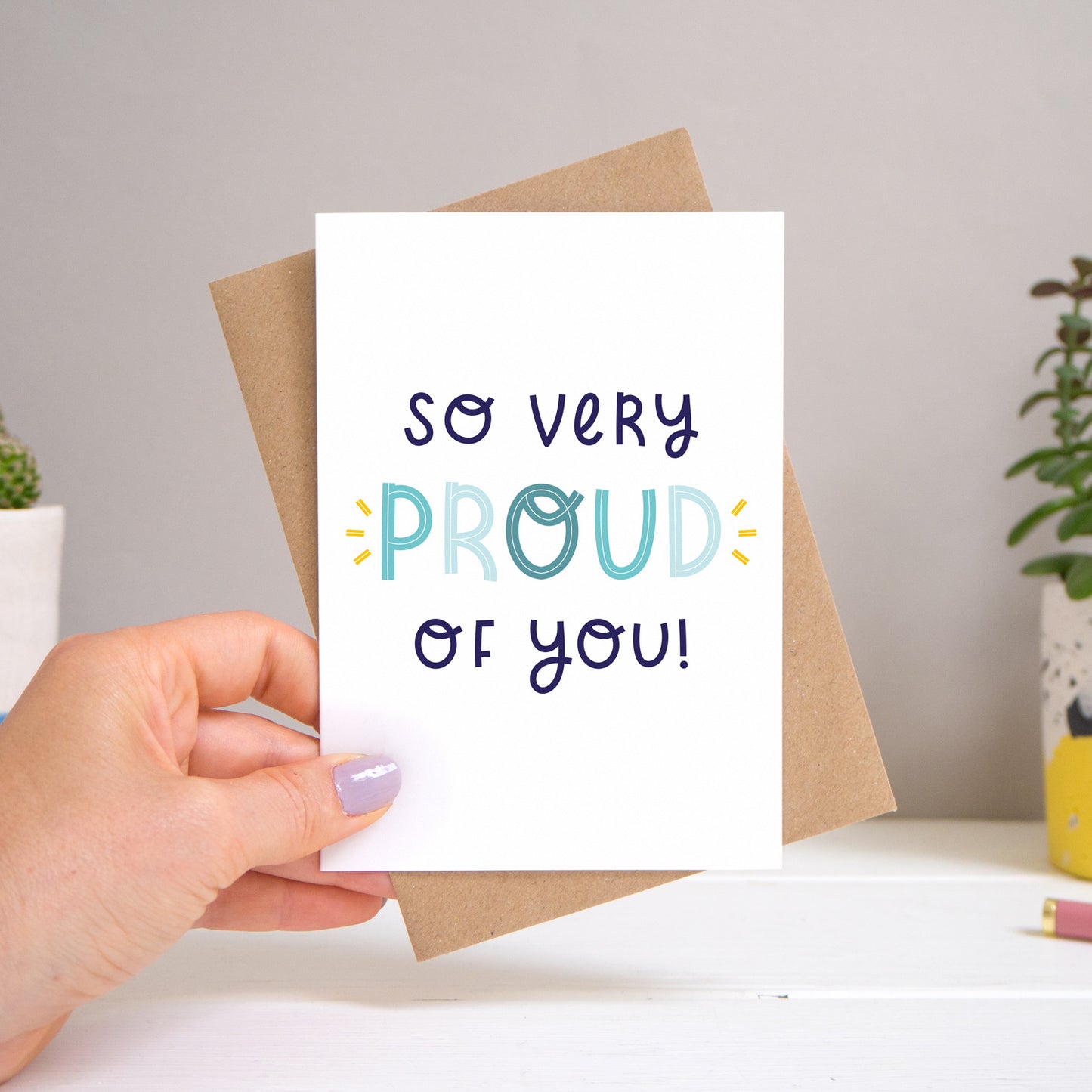 A ‘so very proud of you’ card held over a warm grey and white background with potted plants peeping the sides. Behind the card is a kraft brown envelope that comes with the card. The text on this version of the card is in varying tones of navy and blue.