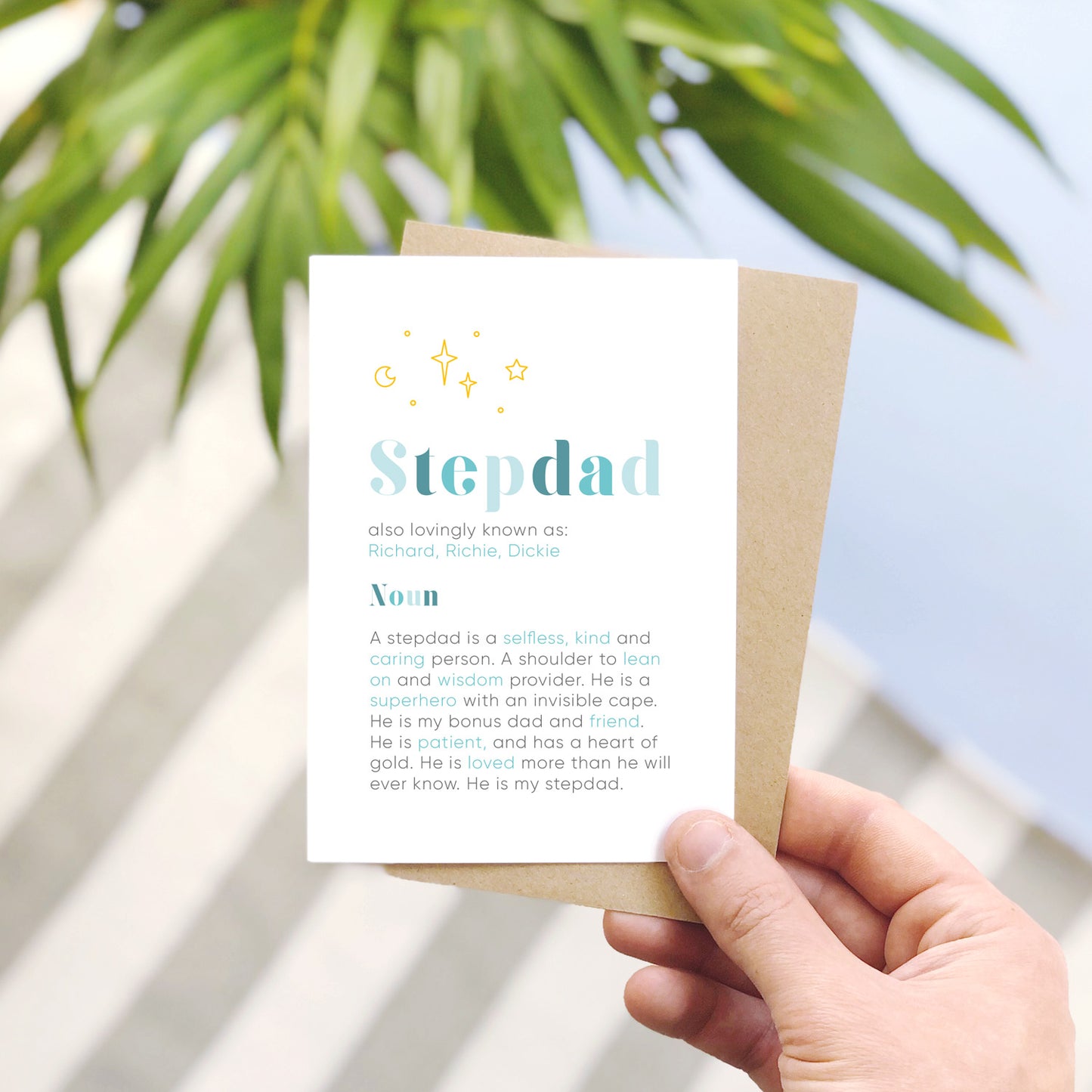 A personalised stepdad definition card being held over a stripy carpet, an area of blue and with a pot plant in the background. Each card has space for nicknames under the blue stepdad text and below features the definition of what it means to be a stepdad.