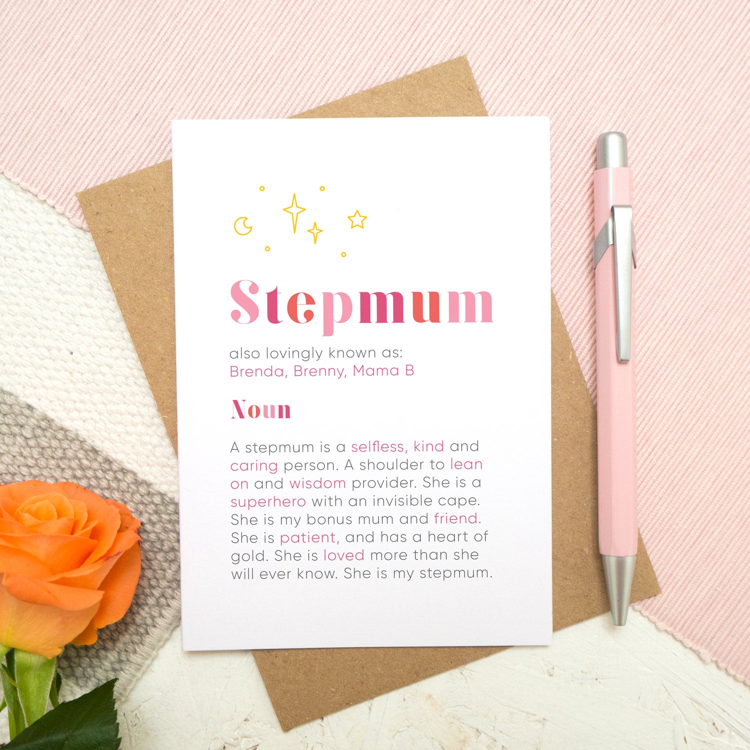 A personalised Stepmum dictionary definition card lying flat on top of a kraft brown envelope on top of a pink runner and a grey and white striped rug. There is an orange rose in the bottom left corner and a pen for scale on the right. The card features hand drawn writing in varying tones of pink and a definition of what a Stepmum is with key words highlighted in pink.