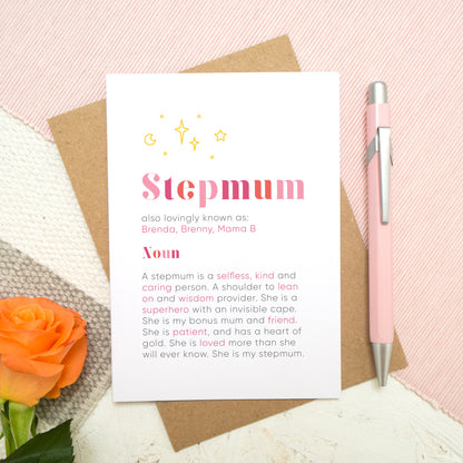 A personalised Stepmum dictionary definition card lying flat on top of a kraft brown envelope on top of a pink runner and a grey and white striped rug. There is an orange rose in the bottom left corner and a pen for scale on the right. The card features hand drawn writing in varying tones of pink and a definition of what a Stepmum is with key words highlighted in pink.