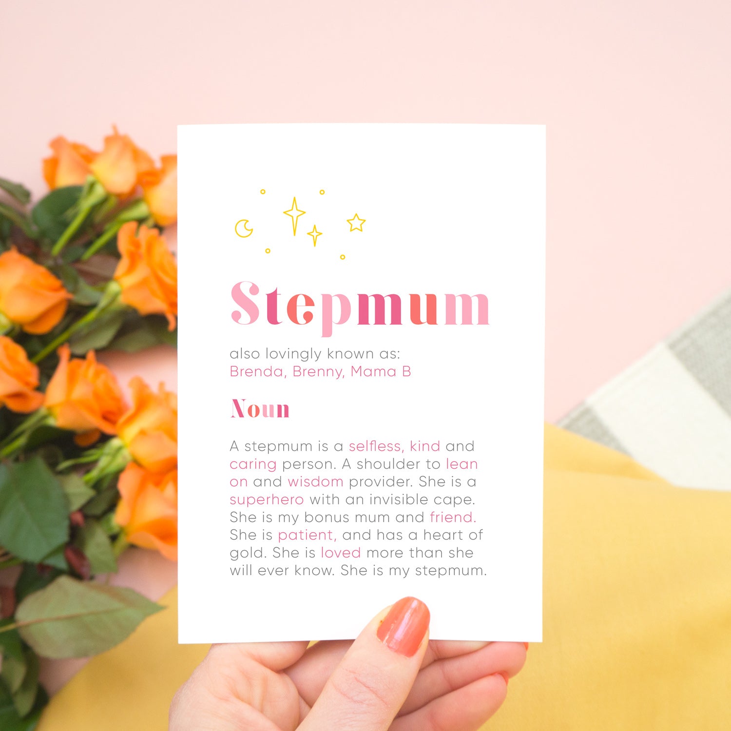 A personalised Stepmum dictionary definition card being held over a pink background with a grey and white striped rug, yellow skirt and a bunch of orange roses to the left. The card features hand drawn writing in varying tones of pink and a definition of what a Stepmum is with key words highlighted in pink.