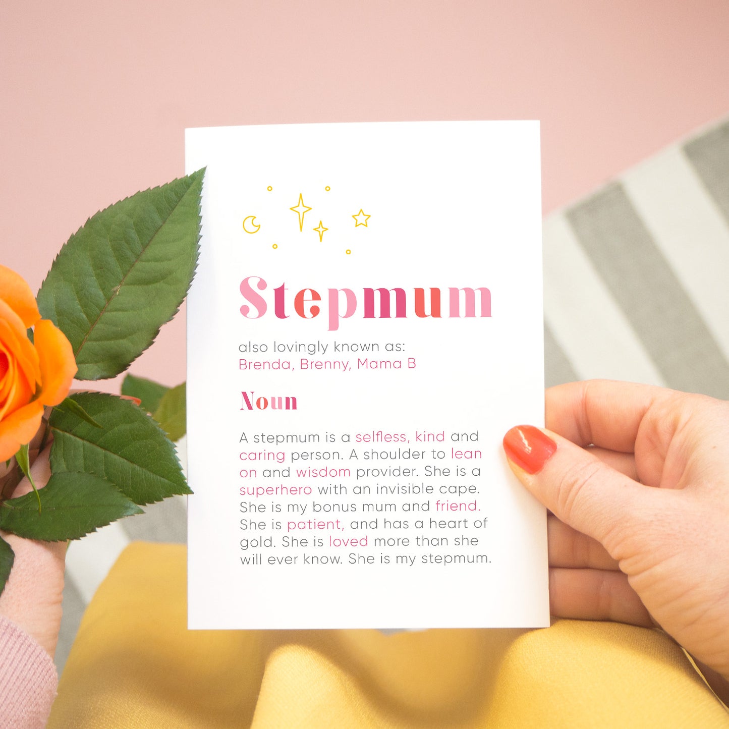 A personalised Stepmum dictionary definition card being held over a pink background with a grey and white striped rug, yellow skirt and a single orange rose to the left. The card features hand drawn writing in varying tones of pink and a definition of what a Stepmum is with key words highlighted in pink.