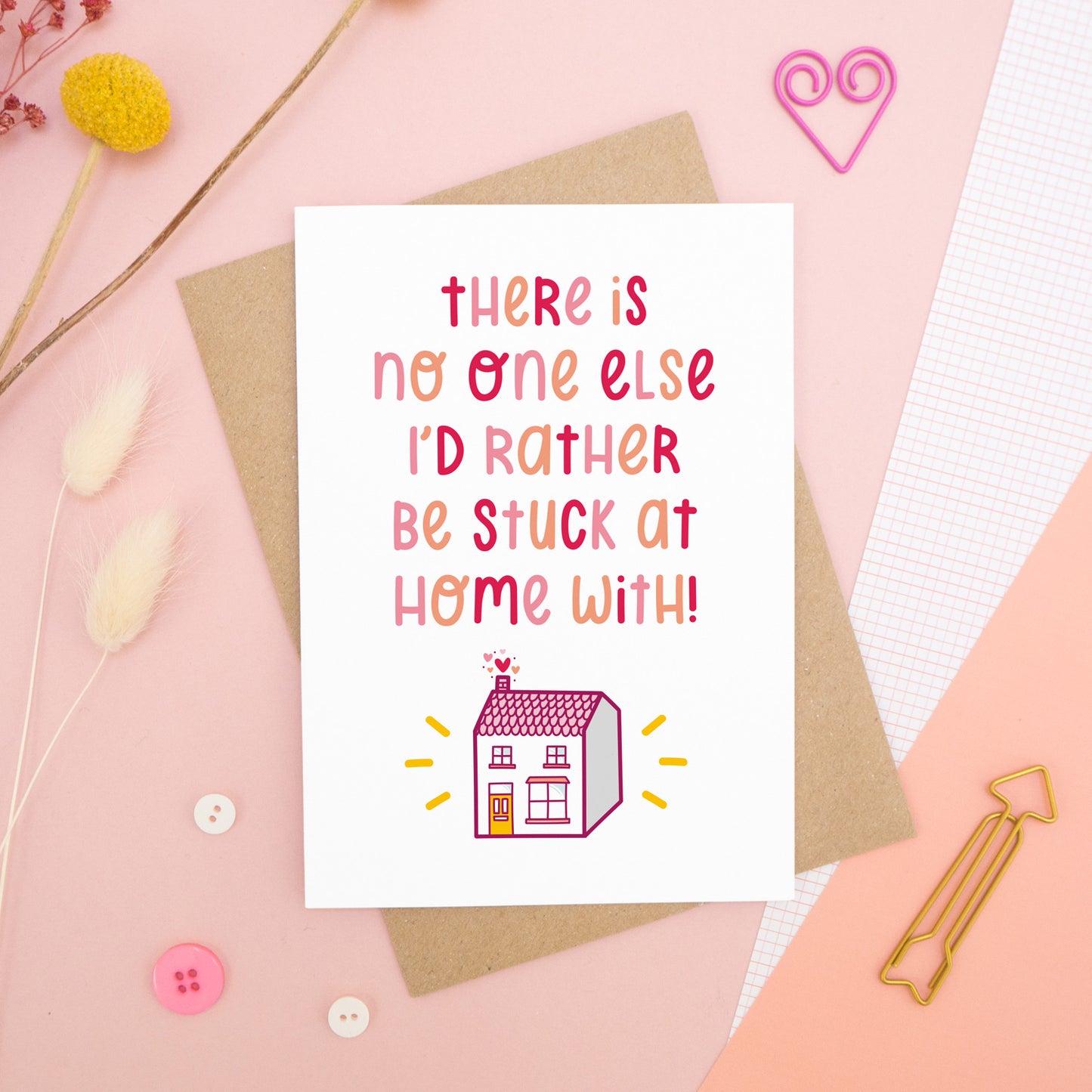 The 'stuck at home with you' card photographed on a pink background with dried flowers, buttons and paper clips as props.