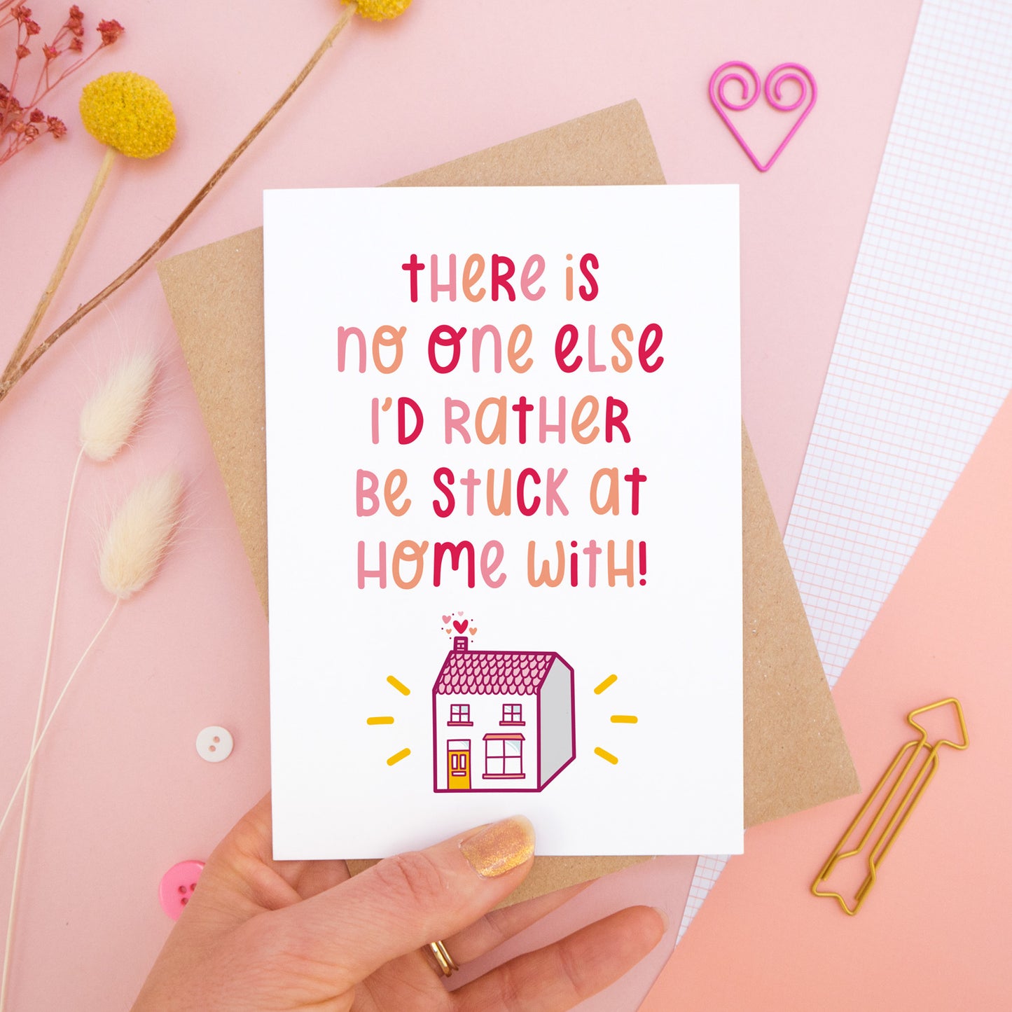 The 'stuck at home with you' card photographed on a pink background with dried flowers, buttons and paper clips as props. The card itself is being held above the scene.
