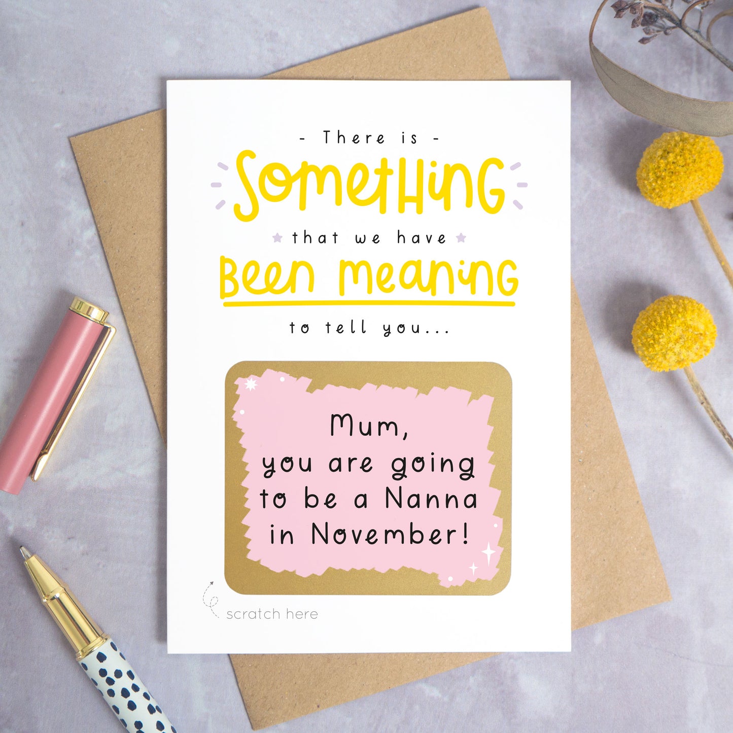 A personalised custom announcement scratch card photographed on a grey background with yellow flowers and a pink pen lid and spotty pen. The card is lying on a kraft brown envelope and the scratch box has been scratched off to reveal the personalised message. This is the yellow and pink colour way.