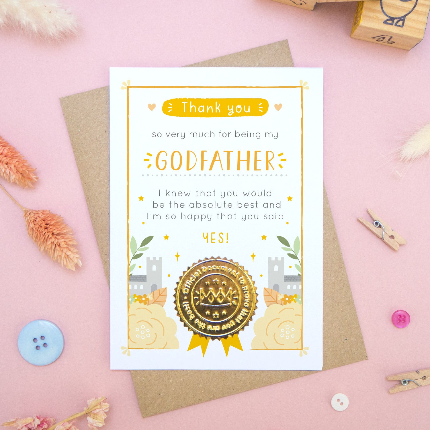 A ‘thank you for being my godfather’ certificate card with a shiny gold seal in the orange colour palette lying flatlay on top of a kraft brown envelope on a pink surface surrounded by building blocks, buttons and dry flowers.