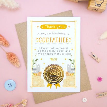 A ‘thank you for being my godfather’ certificate card with a shiny gold seal in the orange colour palette lying flatlay on top of a kraft brown envelope on a pink surface surrounded by building blocks, buttons and dry flowers.