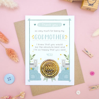 A ‘thank you for being my godmother’ certificate card with a shiny gold seal in the blue colour palette lying flatlay on top of a kraft brown envelope on a pink surface surrounded by building blocks, buttons and dry flowers.