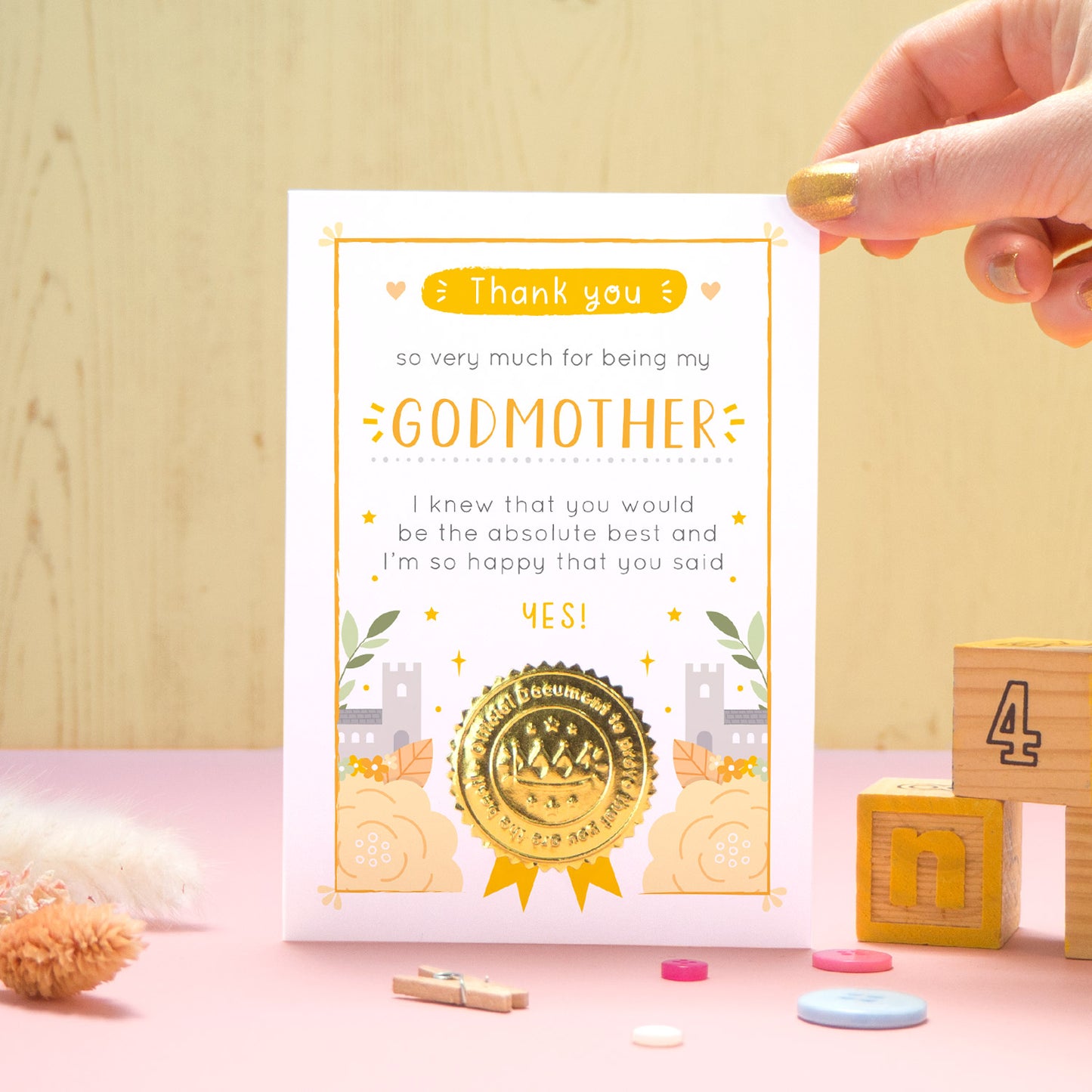 A ‘thank you for being my godmother’ certificate card with a shiny gold seal in the orange colour palette standing in front of a yellow and pink background with building blocks, buttons and dry flowers.