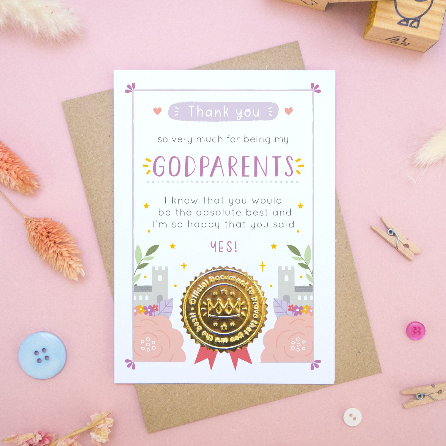 A ‘thank you for being my godparents’ certificate card with a shiny gold seal in the purple colour palette lying flatlay on top of a kraft brown envelope on a pink surface surrounded by building blocks, buttons and dry flowers.