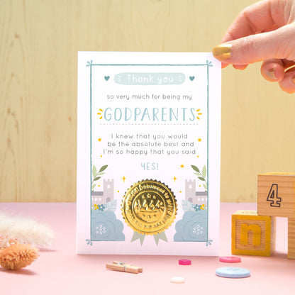 A ‘thank you for being my godparents’ certificate card with a shiny gold seal in the blue colour palette standing in front of a yellow and pink background with building blocks, buttons and dry flowers.