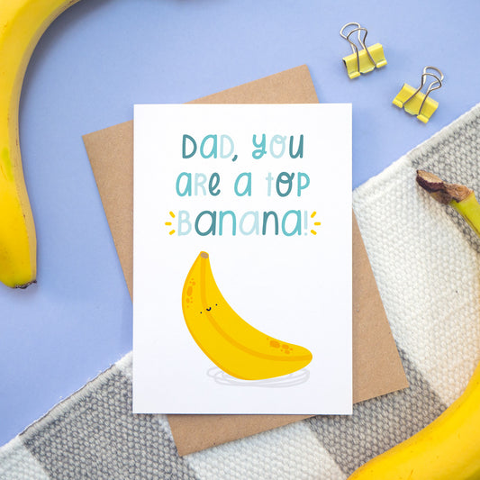 A top banana card photographed on a stripy white and grey rug, with a blue background and surrounded by bananas! The card is lying on a kraft brown envelope and feature blue lettering with a happy bright yellow banana!