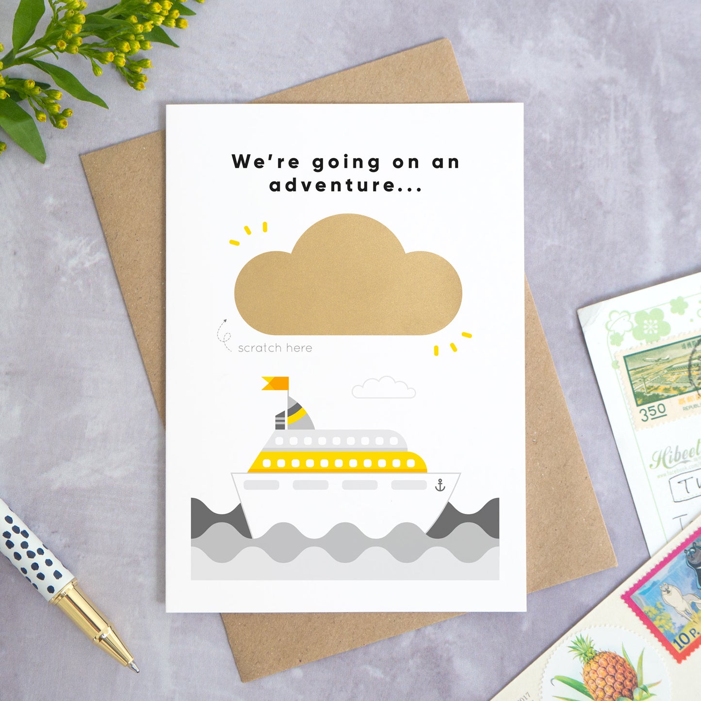 A personalised cruise ship holiday announcement scratch card photographed on a grey concrete background with greenery, a pen and postcards around the edge. The card is set upon a kraft brown envelope. The card is in the grey colour palette and the cloud has yet to be scratched off.
