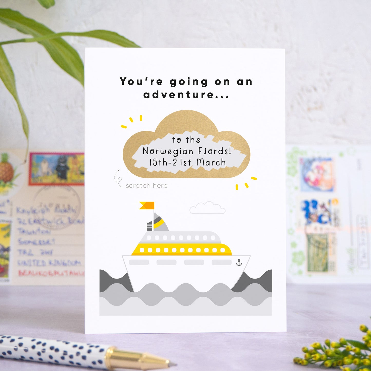 A personalised cruise ship holiday announcement scratch card in the colour grey. The gold cloud has been scratch off to reveal custom holiday details. The card has been shot on a grey and white background with postcards, a pen and greenery in both the fore and background.