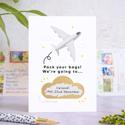 A personalised aeroplane holiday announcement scratch card in the colour grey. The gold cloud has been scratch off to reveal custom holiday details. The card has been shot on a grey and white background with postcards, a pen and greenery in both the fore and background.