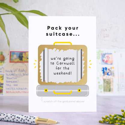 A personalised suitcase travel plans reveal scratch card shot on a grey concrete surface surrounded by greenery, a pen and post cards. The card is the grey version and shows an example message that has been scratched off.