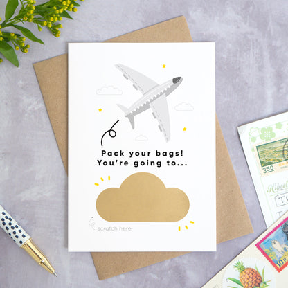 A personalised aeroplane holiday announcement scratch card photographed on a grey concrete background with greenery, a pen and postcards around the edge. The card is set upon a kraft brown envelope. The card is in the grey colour palette and the cloud has not yet been scratched off.