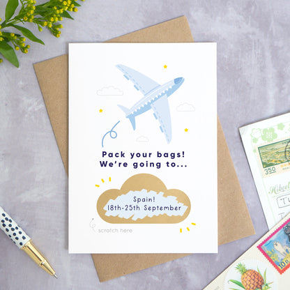 A personalised aeroplane holiday announcement scratch card photographed on a grey concrete background with greenery, a pen and postcards around the edge. The card is set upon a kraft brown envelope. The card is in the blue colour palette and the cloud has been scratched off to reveal a custom message.
