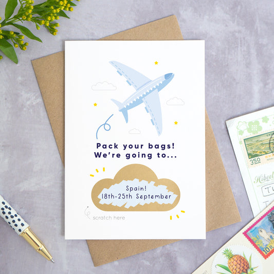 A personalised aeroplane holiday announcement scratch card photographed on a grey concrete background with greenery, a pen and postcards around the edge. The card is set upon a kraft brown envelope. The card is in the blue colour palette and the cloud has been scratched off to reveal a custom message.