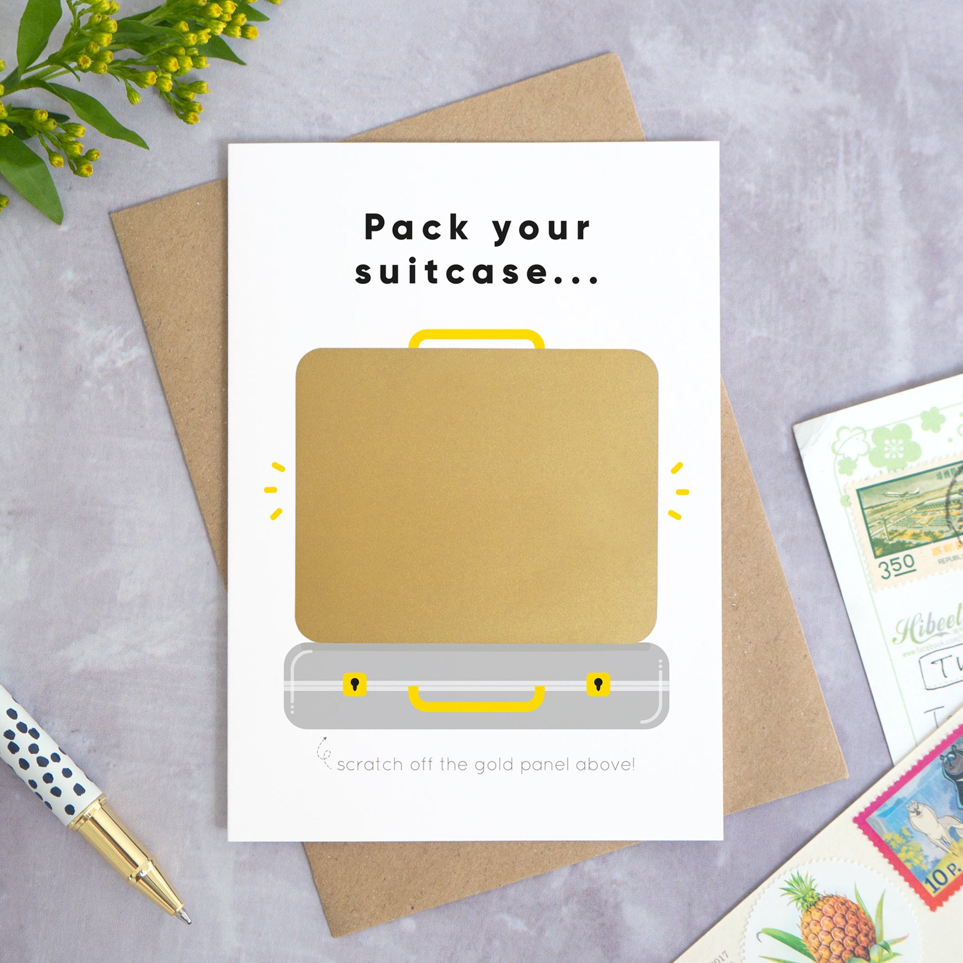 A personalised suitcase travel plans reveal scratch card shot on a grey concrete surface surrounded by greenery, a pen and post cards. The card is the grey version and shows how the card will arrive with the gold panel attached.