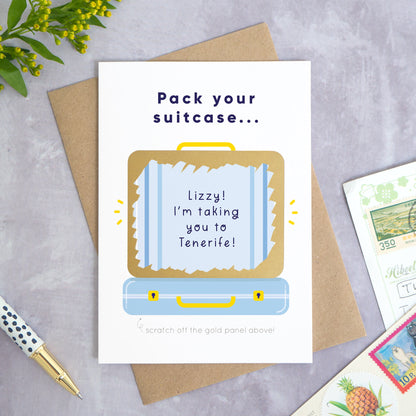 A personalised suitcase travel plans reveal scratch card shot on a grey concrete surface surrounded by greenery, a pen and post cards. The card is the blue version and shows an example message that has been scratched off.