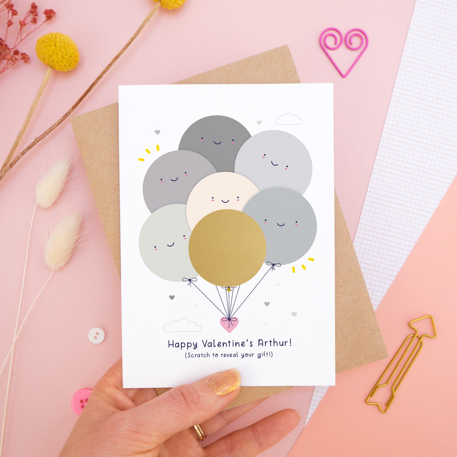 A personalised Valentine’s scratch card photographed on a pink background with floral props, paper clips, and buttons. This card shows the grey colour scheme and the golden circle has yet to be scratched off to reveal the secret message!