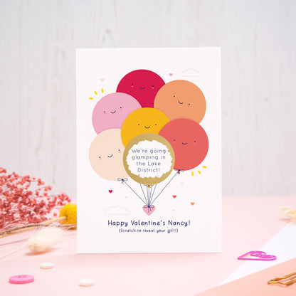 A personalised Valentine’s scratch card photographed on a pink and white background with floral props, paper clips, and buttons. This card shows the red & peach colour scheme and the golden circle has been scratched off to reveal the secret message!